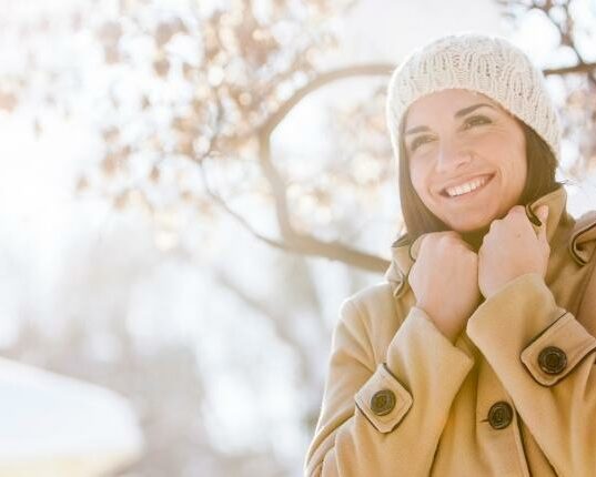 hiver-soins-institut Jeune femme avec un manteau et un bonnet, extérieur enneigé avec du soleil.