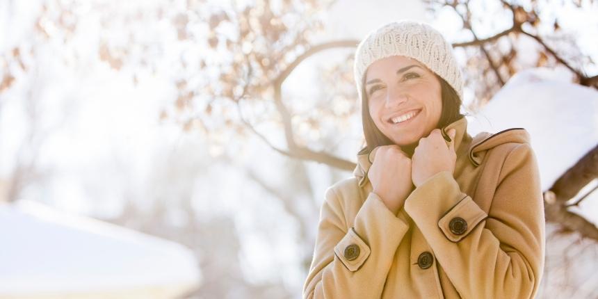 hiver-soins-institut Jeune femme avec un manteau et un bonnet, extérieur enneigé avec du soleil.